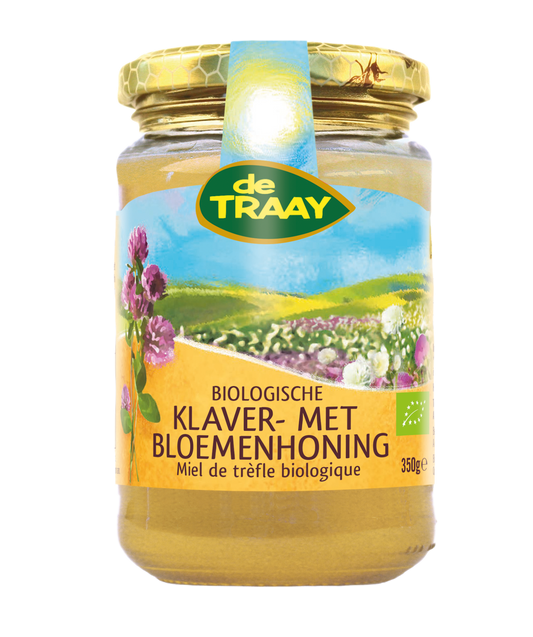 Jar of de Traay organic flower honey, 350g, with honeycomb and flowers visible in the clear glass container.